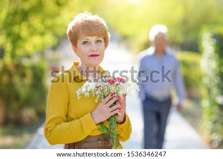An elderly woman is waiting for her beloved man to approach her. A woman with flowers is waiting for her husband.