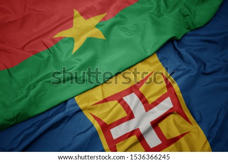 waving colorful flag of madeira and national flag of burkina faso. macro