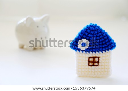 Mini blue roof crocheted house with blurred white ceramic piggy bank on white background. Concept for financial home loan or money saving for house buying