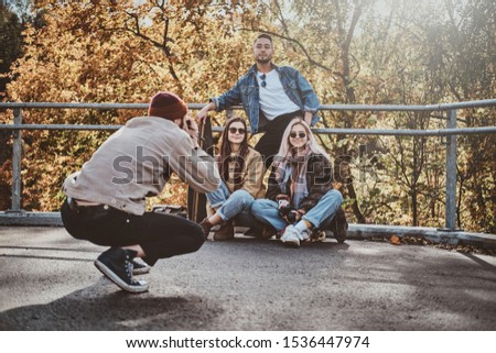 Young man in hat is making a photo of his friends at sunny autumn park.