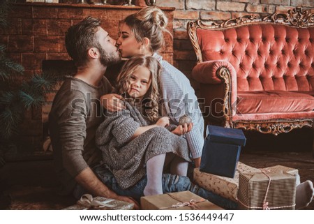 Surrounded by christmas gifts, mother and father are kissing, while holding their little daughter.