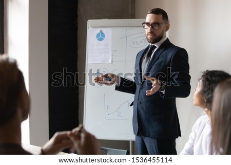 Confident male business trainer coach leader give flip chart presentation at office training, businessman conference speaker wear suit teach executive group explain strategy at corporate workshop Royalty-Free Stock Photo #1536455114
