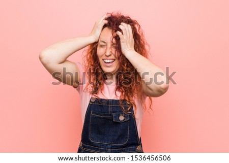 Young pretty ginger redhead woman wearing a jeans dungaree laughs joyfully keeping hands on head. Happiness concept.