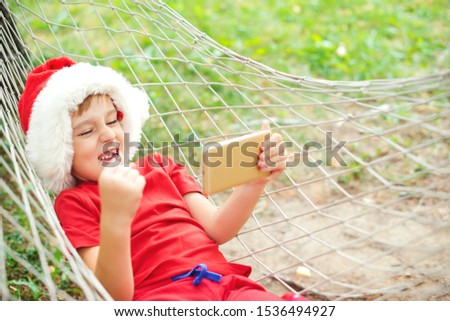 Happy child in Santa hat screaming yes! Excited boy lying in hammock and playing online games. Happy Christmas holidays. Christmas sales, discounts and shopping concept