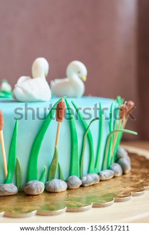 Children's colorful blue fondant birthday cake decorated with little swans and boat on the pond and green reeds at the side of the cake