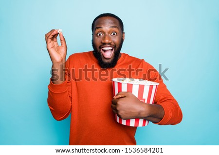 Photo of funny atonished mixed-race crazy ecstatic screaming man watching television series and amazed about plot holding popcorn bucket smiling toothily isolated blue vivid color background