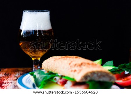 
Selective focus of a glass of beer behind a sandwich of raw ham, avocado, cheese, tomato and arugula, on a wooden table with a black background.