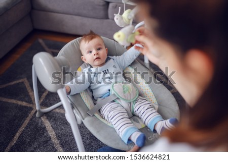 Adorable baby boy lying in bouncer an looking at his mother. Mother entertaining her loving son. Living room interior. Royalty-Free Stock Photo #1536624821