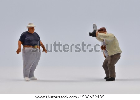 Miniature  photographer is taking a picture of a corpulent guy  with a hat on his head