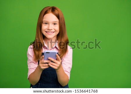 Photo of cheerful pretty little schoolchild holding telephone hands texting with classmates friends wear denim overall pink shirt isolated green background