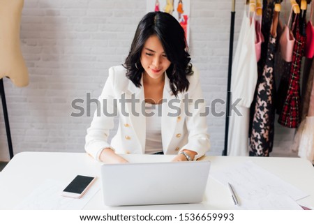 Contemporary confident Asian lady entrepreneur in stylish jacket working on laptop with garment hanger on background