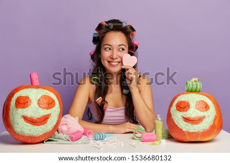 Smiling beautiful multiracial female uses makeup products, holds cosmetic sponge for face care, has long hair with curlers, sits against purple background, tries new cream on orange pumpkins