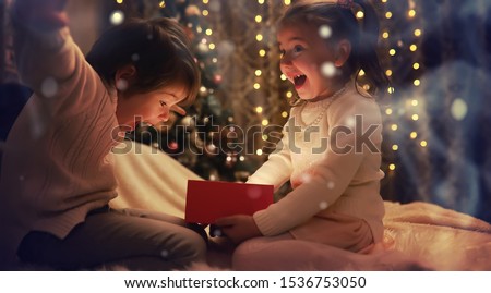 Family on Christmas eve at fireplace. Kids opening Xmas presents. Children under Christmas tree with gift boxes. Decorated living room with traditional fire place. Cozy warm winter evening home.