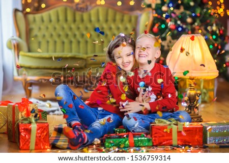 Happy children are sitting on the floor near gifts under the falling candy and hugging, against the background of a Christmas tree and holiday lights. Cristmas presents.