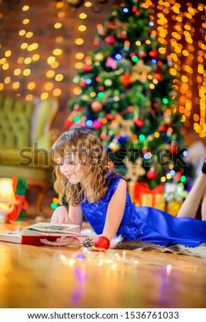 A girl in a blue dress lying on the floor in the room reading a book and smiling, against the background of a festive tree. Christmas time.