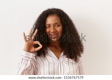Everything is perfect. Positive pretty girl of African origin posing against white blank wall background, winking at camera and making okay gesture, connecting thumb and index finger. Body language