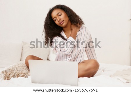 Stylish young dark skinned female copywriter working distantly from home sitting on bed in silk pajamas in front of open electronic gadget, editing her new article, checking it for mistakes