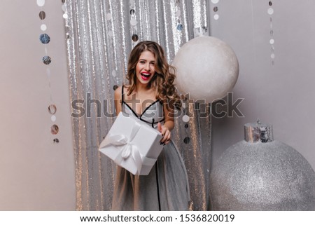 Girl with long dark hair and beautiful make-up in good mood at Christmas party. Portrait of lady who received holiday gift in beautifully decorated studio