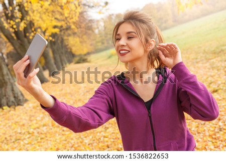 Young woman standing outdoors in the autumn park taking selfie photos on smartphone posing to camera smiling cheerful close-up