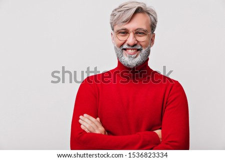 Laughing old man with white beard posing with arms crossed. Indoor photo of amazing male model in red sweater.