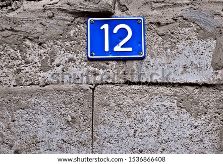 Number 12, twelve, blue plate on a gray rough concrete surface.