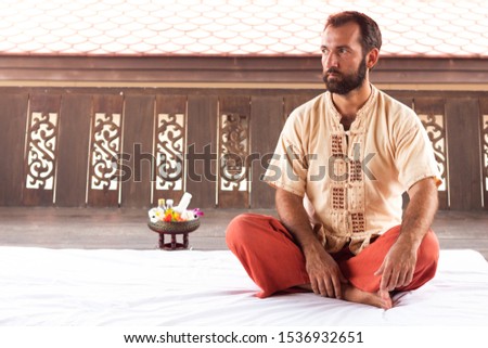 Thai massage teacher and therapist ready to teach students traditional nuad boran techniques. Man sit on white yoga mat on red trouses. Flower and instruments on background. Yoga coaching instructor. Royalty-Free Stock Photo #1536932651
