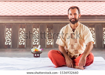Thai massage teacher and therapist ready to teach students traditional nuad boran techniques. Man sit on white yoga mat on red trouses. Flower and instruments on background. Yoga coaching instructor. Royalty-Free Stock Photo #1536932660