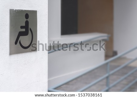 Ragged wheelchair sign or handicapped symbol on a steel plate stick on flat white cement wall with blurred outdoor disable ramp background. 