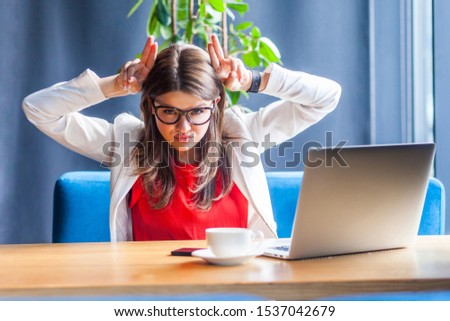 Portrait of aggressive beautiful stylish brunette young woman in glasses sitting and looking at camera with cow horns on head and serious anger face. indoor studio shot, cafe, office background.
