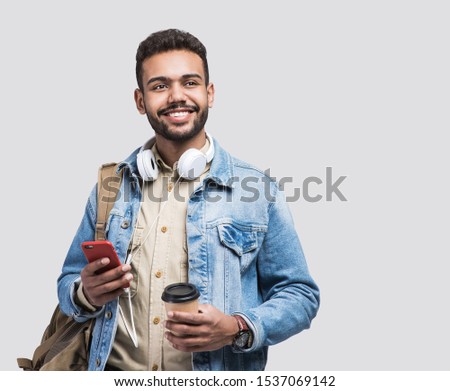 Young handsome man with backpack holding smart phone and coffee. Smiling student going on travel. Isolated on gray background 