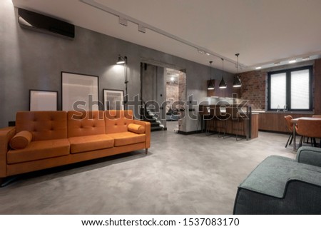 Modern spacious interior of studio flat including brown wooden kitchen with dinning furniture sets and soft orange couch with gray armchair against red brick and gray plastered walls in loft style