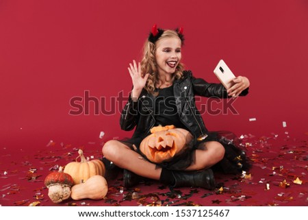 Picture of smiling happy positive little girl devil in carnival halloween costume isolated over red wall background holding pumpkin talking by mobile phone waving.