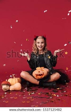 Picture of cheery positive little girl devil in carnival halloween costume isolated over red wall background holding pumpkin over confetti.