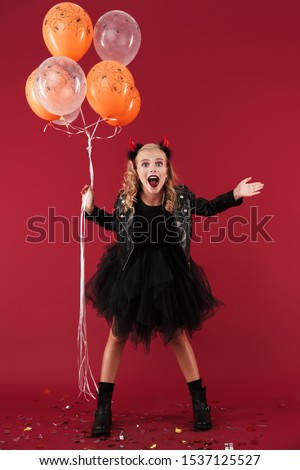 Picture of emotional surprised positive little girl devil in carnival halloween costume isolated over red wall background holding balloons.