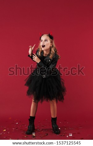 Picture of emotional little girl devil in carnival halloween costume isolated over red wall background.