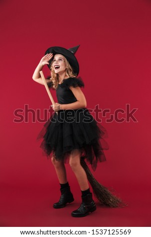 Image of cheery little girl witch in carnival halloween costume isolated over red wall background with broom.