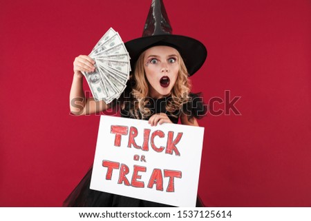Photo of surprised shocked little girl witch in carnival halloween costume isolated over red wall background holding copyspace blank with trick or treat text and money.