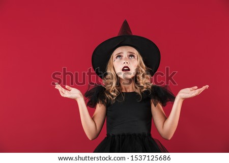 Photo of confused little girl witch in carnival halloween costume isolated over red wall background.