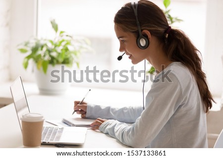 Side view concentrated millennial mixed race woman wearing headset with microphone, looking at laptop screen, listening educational webinar, taking notes, getting remote online education at home. Royalty-Free Stock Photo #1537133861