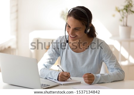 Happy mixed race young woman wearing headset, looking at laptop screen, watching educational lecture, enjoying interesting webinar, writing notes. Smiling employee holding video call with clients. Royalty-Free Stock Photo #1537134842