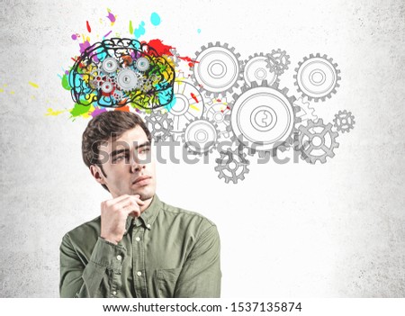 Handsome young man in green shirt standing near concrete wall with colorful brain sketch and gears drawn on it. Concept of brainstorming and creativity