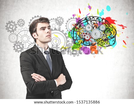 Confident young businessman standing with crossed arms near concrete wall with colorful brain with gears drawn on it. Concept of brainstorming