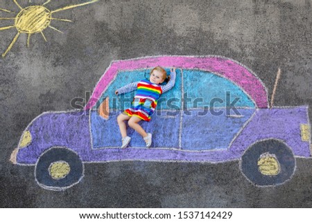 Adorable little toddler girl playing with colorful chalks and painting big car picture on asphalt. Happy baby child playing outside. Creative leisure for children outdoors in summer