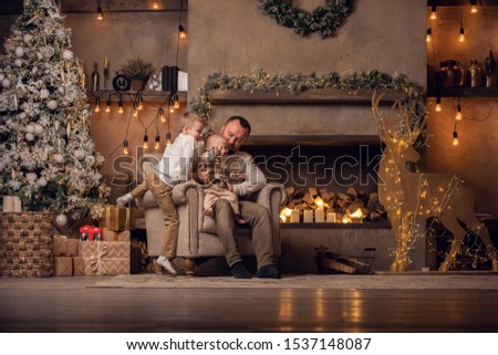 Picture of father with his sons sitting in armchair by New Year tree and fireplace.