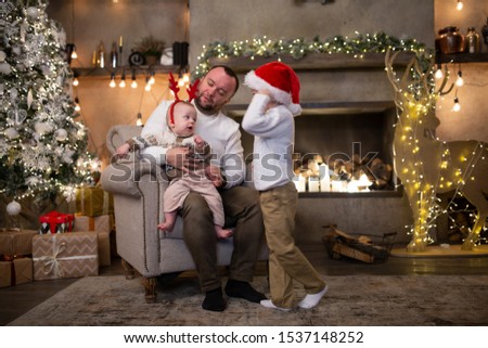 Picture of father with his sons in santa cap sitting in armchair by New Year tree and fireplace