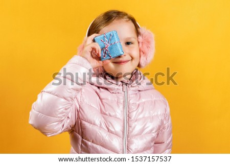 Cheerful little girl with a small box with a gift. A child in a jacket and warm headphones on a yellow background. The concept of Christmas, New Year, winter.