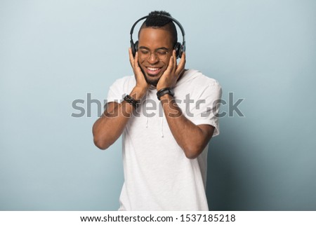 Happy african American millennial man in earphones isolated on blue studio background enjoy music, smiling biracial male in glasses wear modern headphones listen to favorite tracks