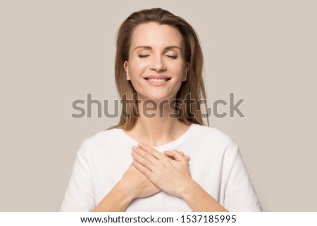 Happy Caucasian young woman in white tshirt isolated on grey studio background hold hands on heart chest feel thankful, smiling grateful millennial female thanking god, show love, empathy and care Royalty-Free Stock Photo #1537185995