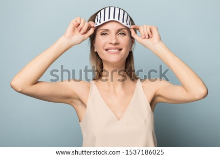 Smiling millennial Caucasian female isolated on blue studio background wearing striped sleeping eye mask on head, happy young woman look at camera pleased with good beauty salon skin procedure result