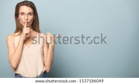 Millennial woman in glasses isolated on blue studio background point at blank copy space make hush gesture, Caucasian female in spectacles share sale promotion offer, look at camera ask be quiet